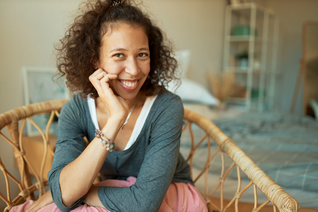 people, rest, relaxation and leisure concept. portrait of young mixed race female wearing casual clothes relaxing in bedroom sitting comfortable in armchair, smiling happily, touching face